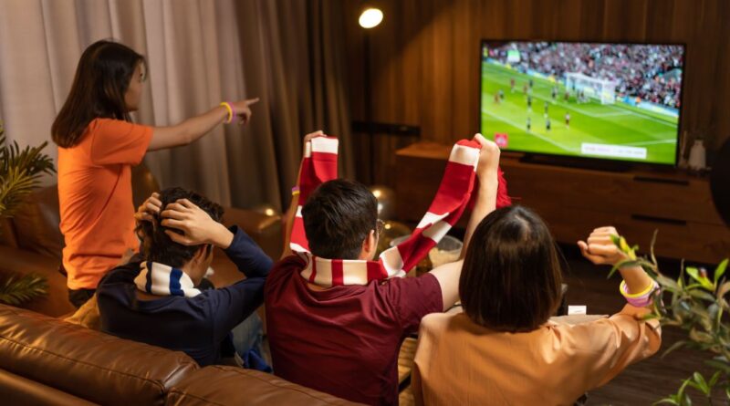 Group of Asian people friends sit on sofa watching and cheering football or soccer games competition on TV together at home